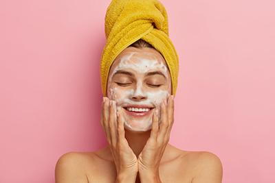 smiling woman eyes closed washing face and yellow towel over hair pink background